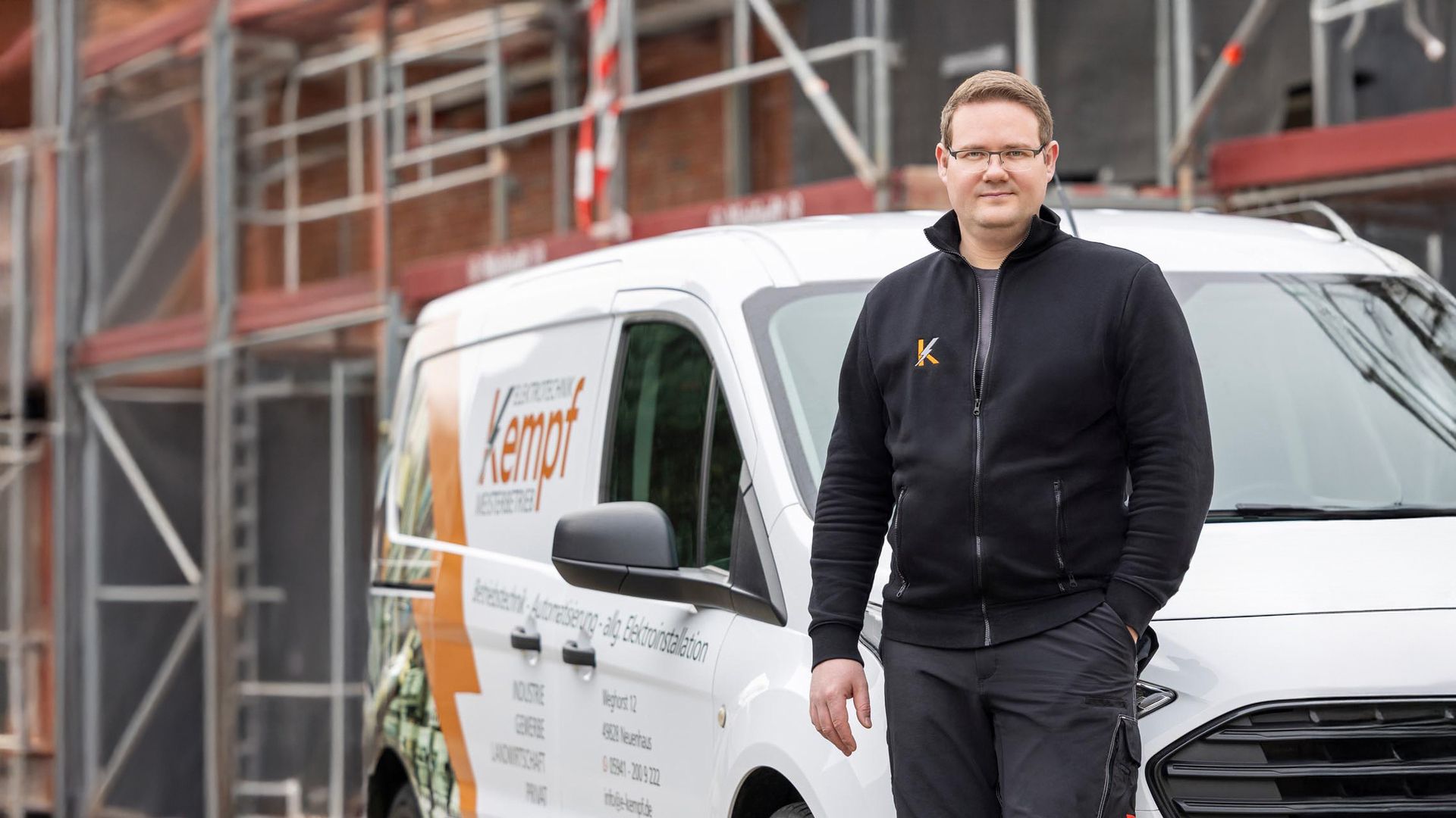 Mann in schwarzer Jacke steht vor weißem Transporter mit orangefarbenem Logo auf einer Baustelle mit Gerüst im Hintergrund